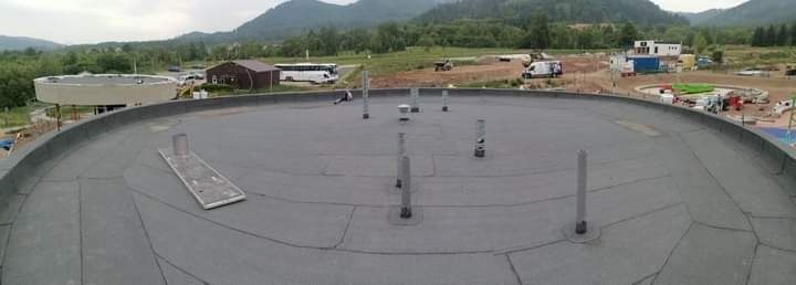 Panoramiczny widok na płaski, okrągły dach pokryty szarą papą z wystającymi pionowymi rurami wentylacyjnymi, w tle zabudowania i zalesione wzgórza.