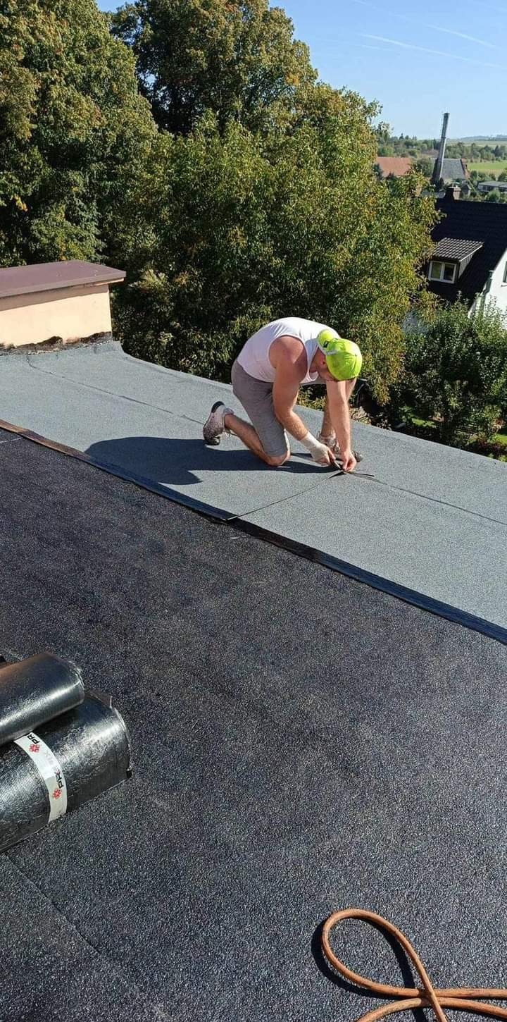 Dekarz w białym podkoszulku i szarych spodenkach, klęczący na dachu pokrytym papą, przycina nożem fragment papy. W tle widoczne drzewa i zabudowania.