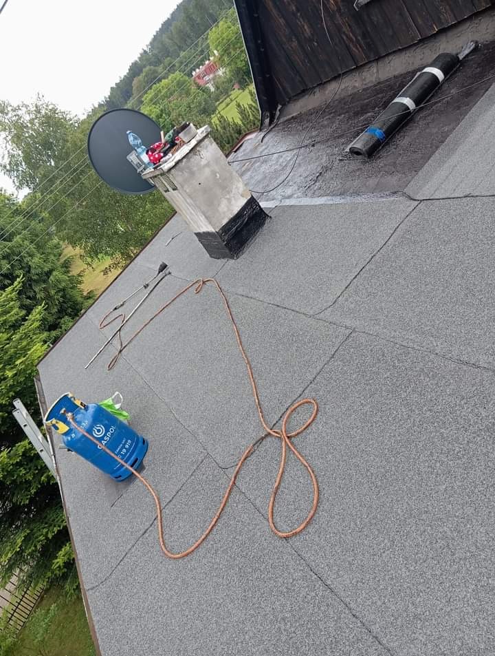 Szary dach pokryty papą termozgrzewalną, widoczny komin z anteną satelitarną, butla gazowa, palnik dekarski i drabina oparta o ścianę, w tle zieleń drzew i budynek.