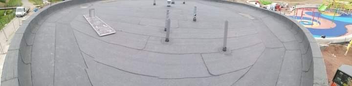 Szeroki kadr na płaski dach pokryty papą, z widocznymi kominkami wentylacyjnymi i elementami mocującymi, w tle plac zabaw i pojazdy budowlane.