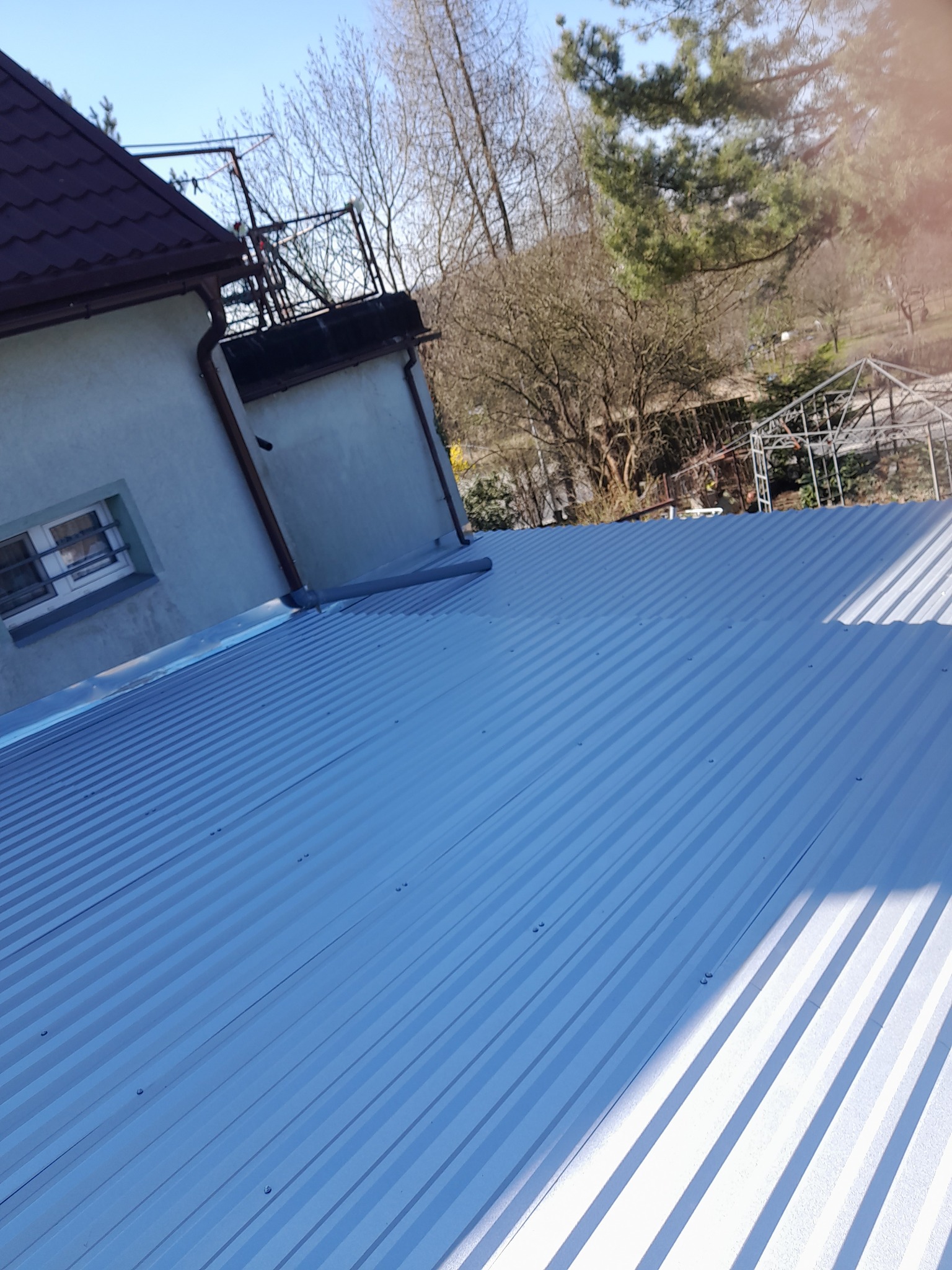 Błękitny dach z blachy trapezowej na jasnym budynku w słoneczny dzień. Widoczne detale mocowań, rynna i fragment ogrodu z konstrukcją szklarni.