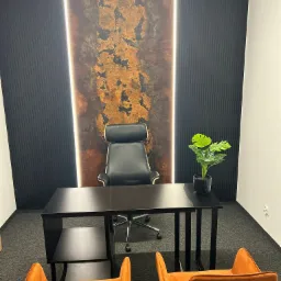 Nowoczesne biuro z czarnym biurkiem, dwoma pomarańczowymi krzesłami, czarnym fotelem biurowym i ścianą z dekoracyjnym panelem podświetlonym pionowymi lampami LED.