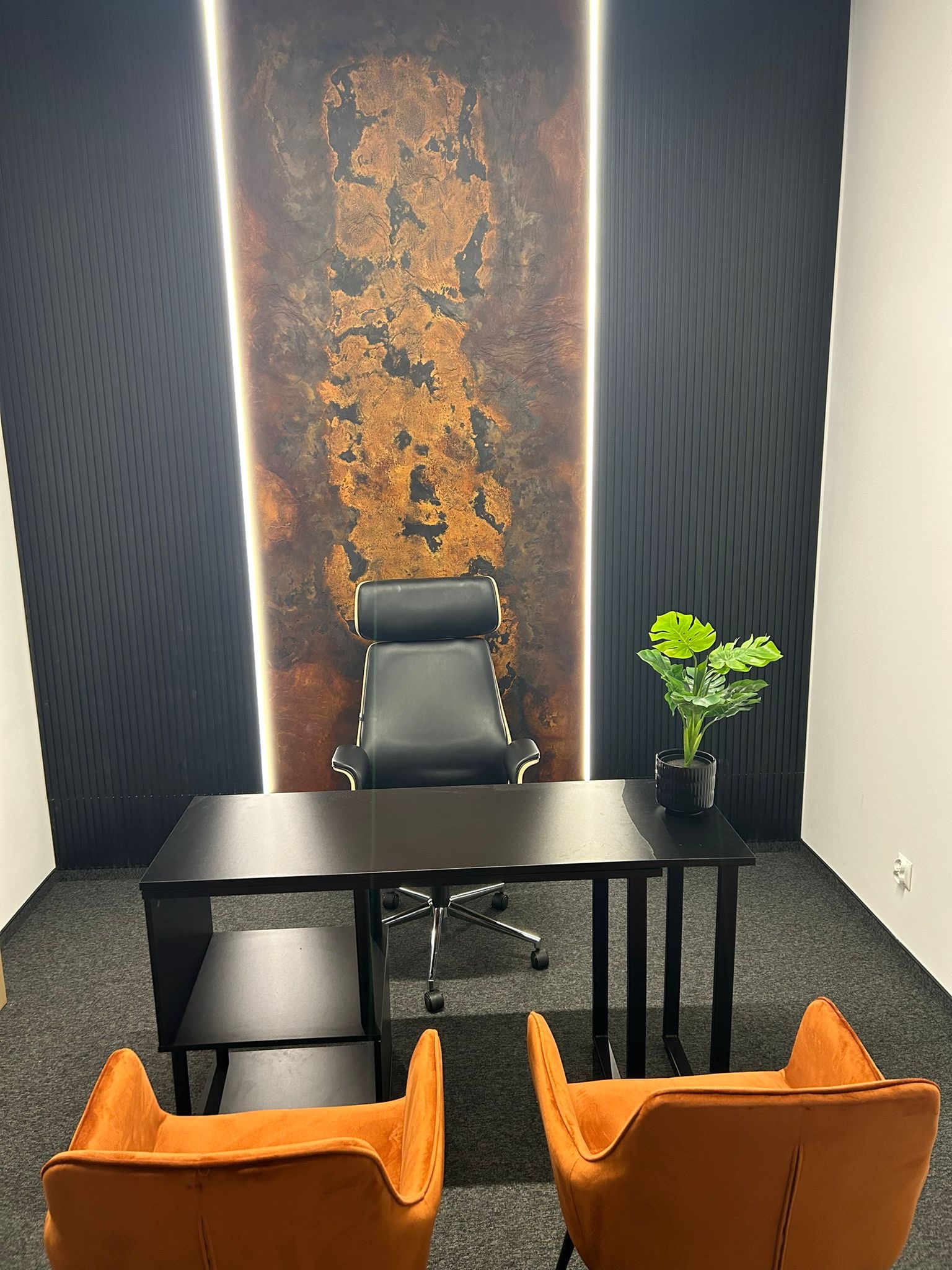 Nowoczesne biuro z czarnym biurkiem, dwoma pomarańczowymi krzesłami, czarnym fotelem biurowym i ścianą z dekoracyjnym panelem podświetlonym pionowymi lampami LED.