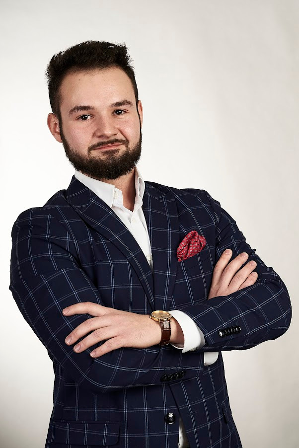 Młody mężczyzna w granatowej marynarce w kratę, białej koszuli i z czerwoną poszetką, z zegarkiem na ręce, pozuje na jasnym tle, z rękami skrzyżowanymi na piersi.