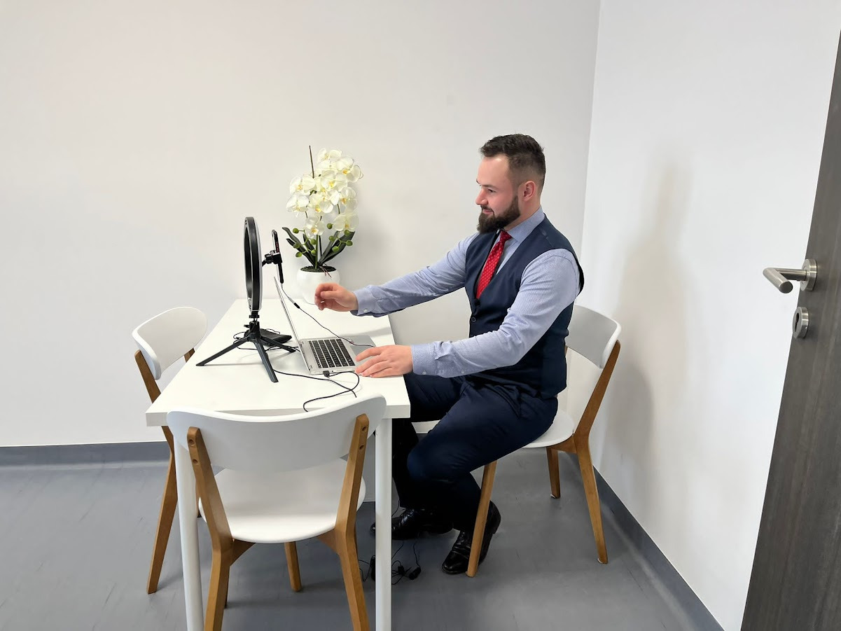 Mężczyzna w garniturze siedzi przy białym stole, korzystając z laptopa i lampy pierścieniowej do wideokonferencji, z kwiatami w tle.
