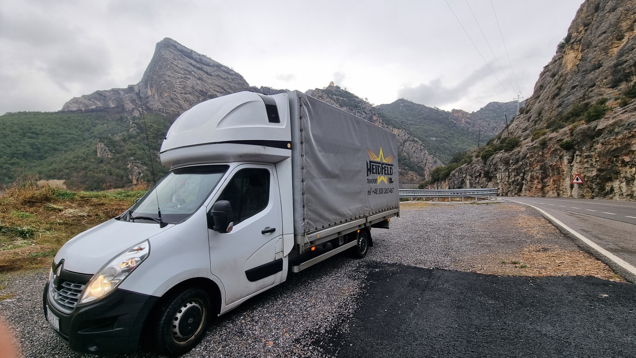 Biały samochód dostawczy z szarą plandeką z logo firmy transportowej na tle górskiego krajobrazu z serpentyną drogi.