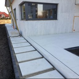 Trivo - Nowoczesny taras z jasnych, betonowych płyt, z obrzeżem z ciemnej kostki, prowadzący do wejścia do domu z widocznym oknem i drzwiami.