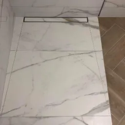 Białe, marmurowe płytki podłogowe w kabinie prysznicowej z liniowym odpływem, widok z góry, fragment drewnopodobnej podłogi w tle.