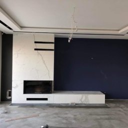 Trivo - Wnętrze w trakcie prac wykończeniowych: ściana akcentowa w kolorze granatowym, zabudowa kominkowa z białego marmuru z czarnymi akcentami, surowa podłoga, widoczne niedokończone instalacje...