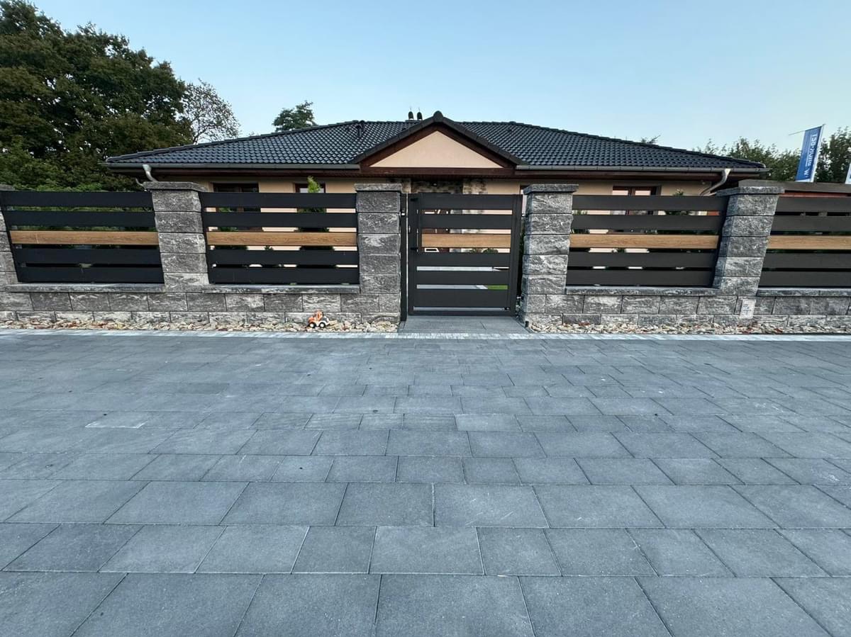 Nowoczesne ogrodzenie posesji z szarej kostki łupanej, czarnych paneli i drewnianych akcentów, z bramą w kolorze antracytu i wybrukowanym podjazdem.