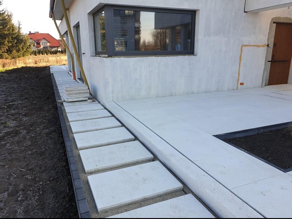 Nowoczesny taras z jasnych, betonowych płyt, z obrzeżem z ciemnej kostki, prowadzący do wejścia do domu z widocznym oknem i drzwiami.