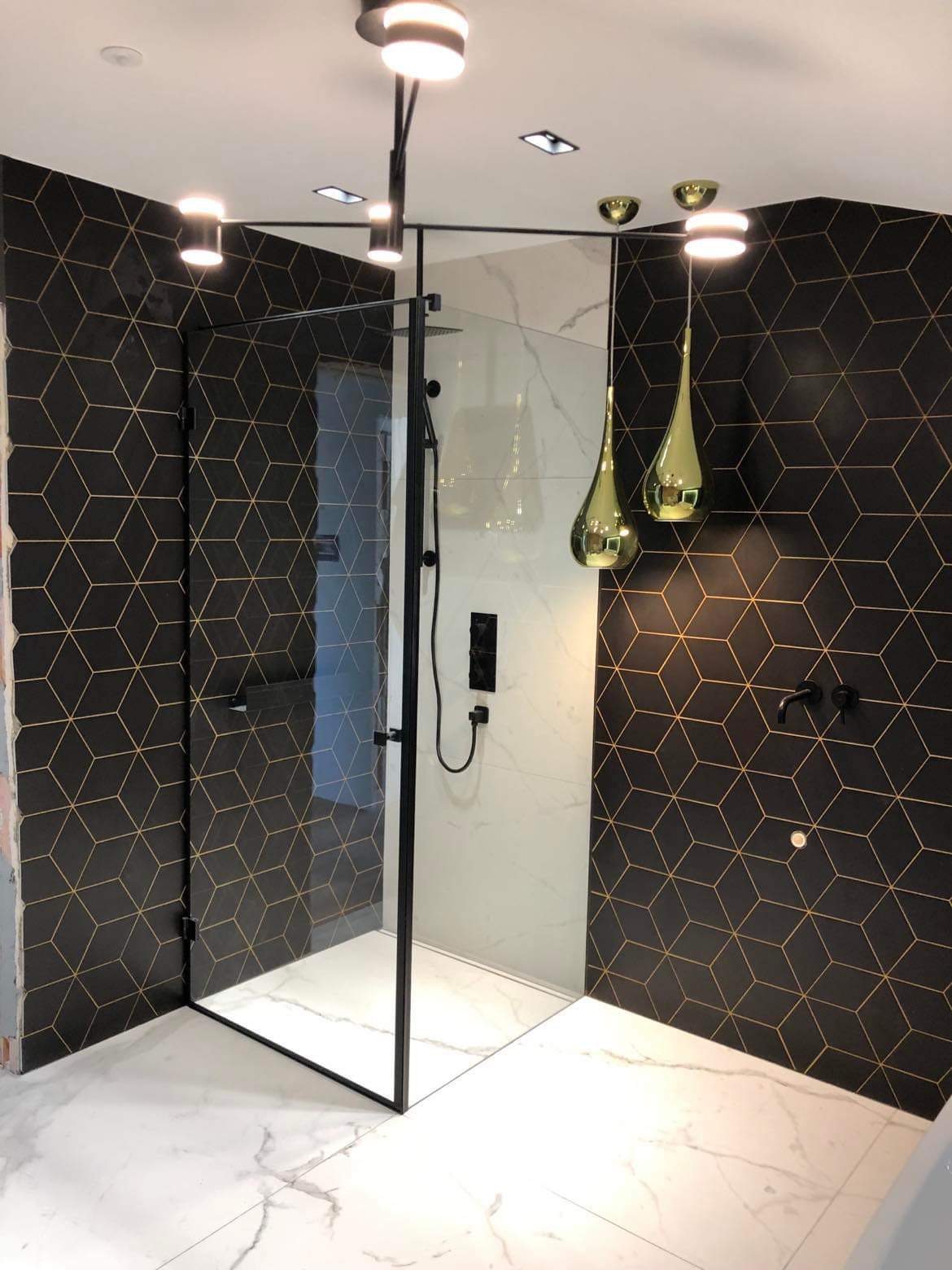Nowoczesna łazienka z czarnymi płytkami w złote geometryczne wzory, kabina prysznicowa ze szklanymi drzwiami i złotymi lampami wiszącymi w kształcie kropli.