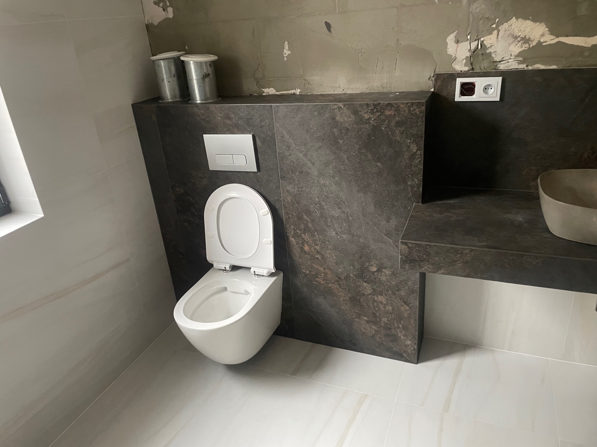 Nowoczesna, podwieszana toaleta z otwartą deską, zamontowana na tle ściany wykończonej ciemnym, marmurkowym gresem, z widocznymi dwoma metalowymi pojemnikami na blacie i gniazdkiem elektrycznym...