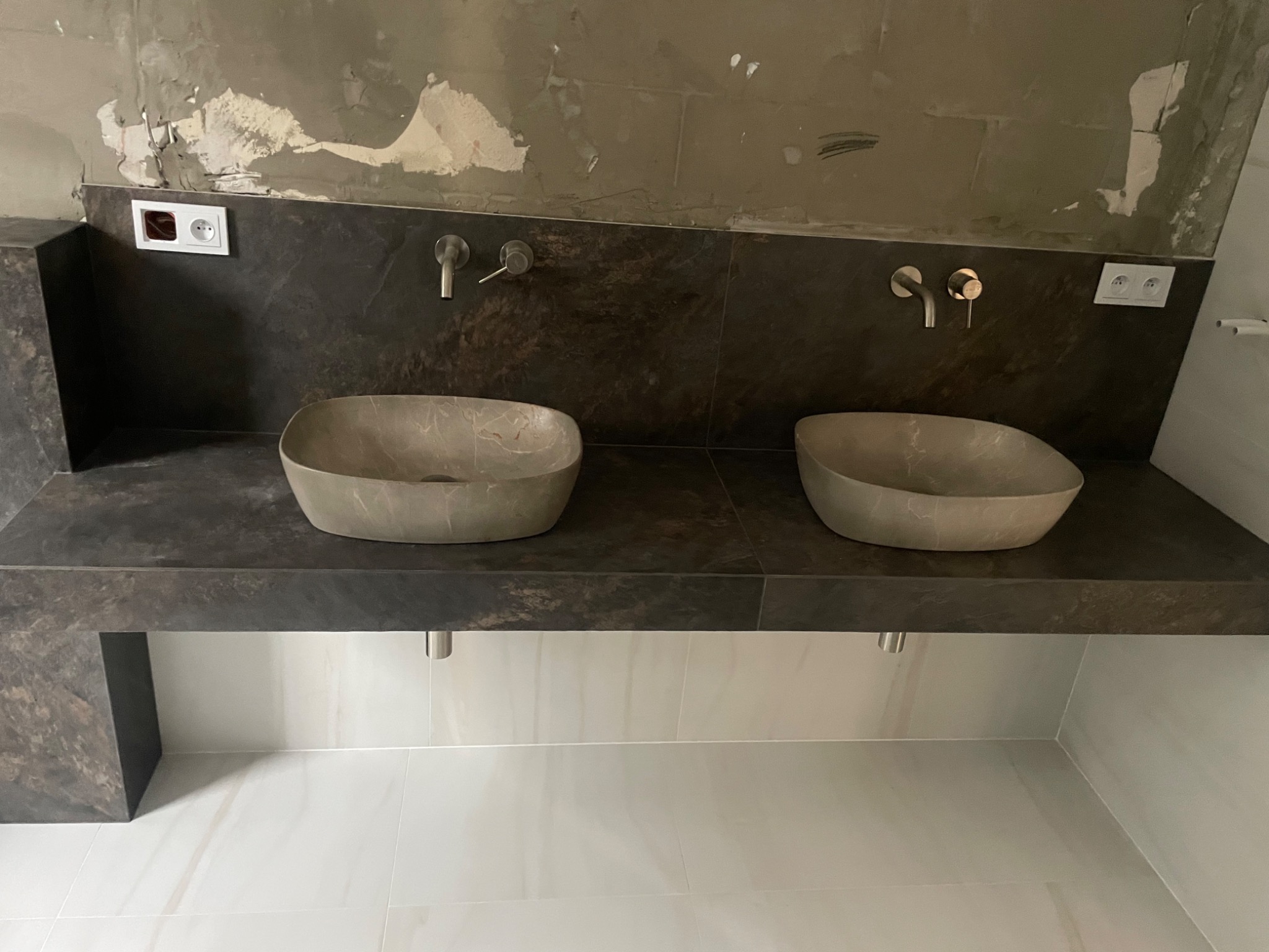 Dwie kamienne umywalki o nieregularnym kształcie na blacie z ciemnego kamienia, widoczne baterie, gniazdka elektryczne i surowa ściana w trakcie prac wykończeniowych.