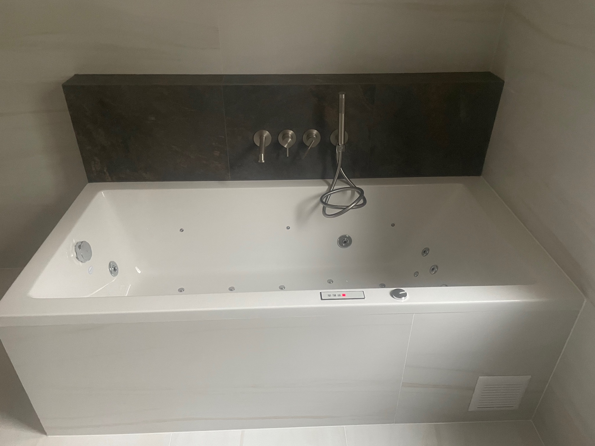 Biała wanna z hydromasażem, chromowana bateria ścienna i dyskretna obudowa z ciemnego kamienia, minimalistyczna łazienka w jasnych płytkach.