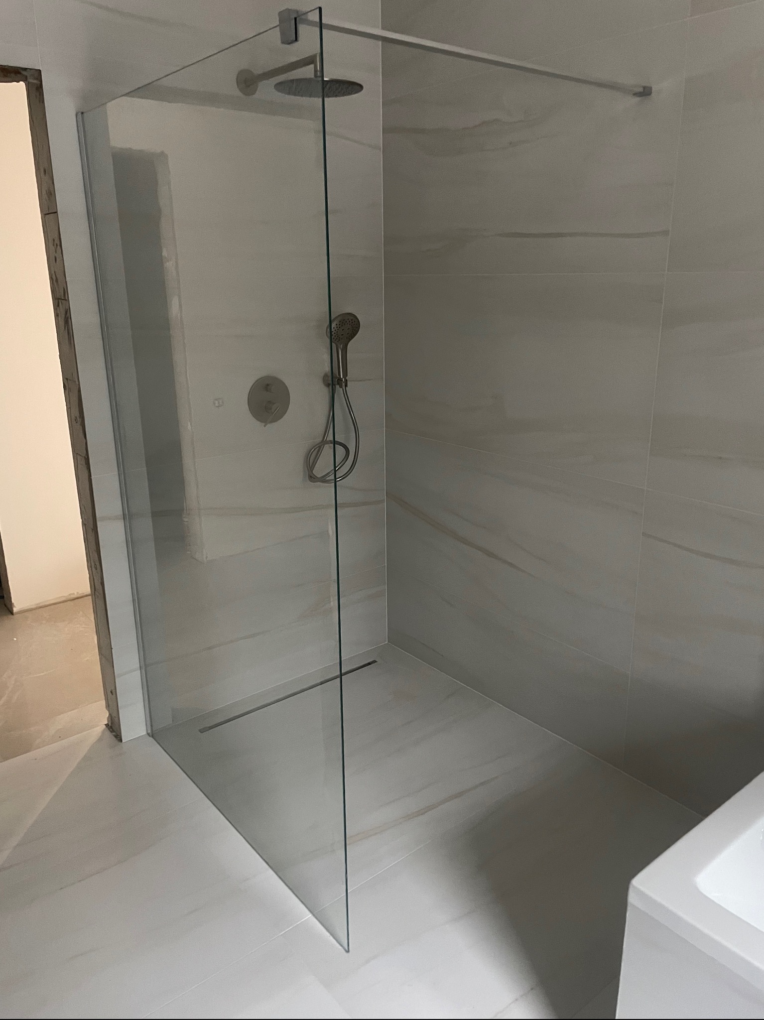 Kabina prysznicowa typu walk-in ze szklaną ścianką, zamontowana w łazience wykończonej płytkami imitującymi marmur, widoczna deszczownica i bateria podtynkowa.