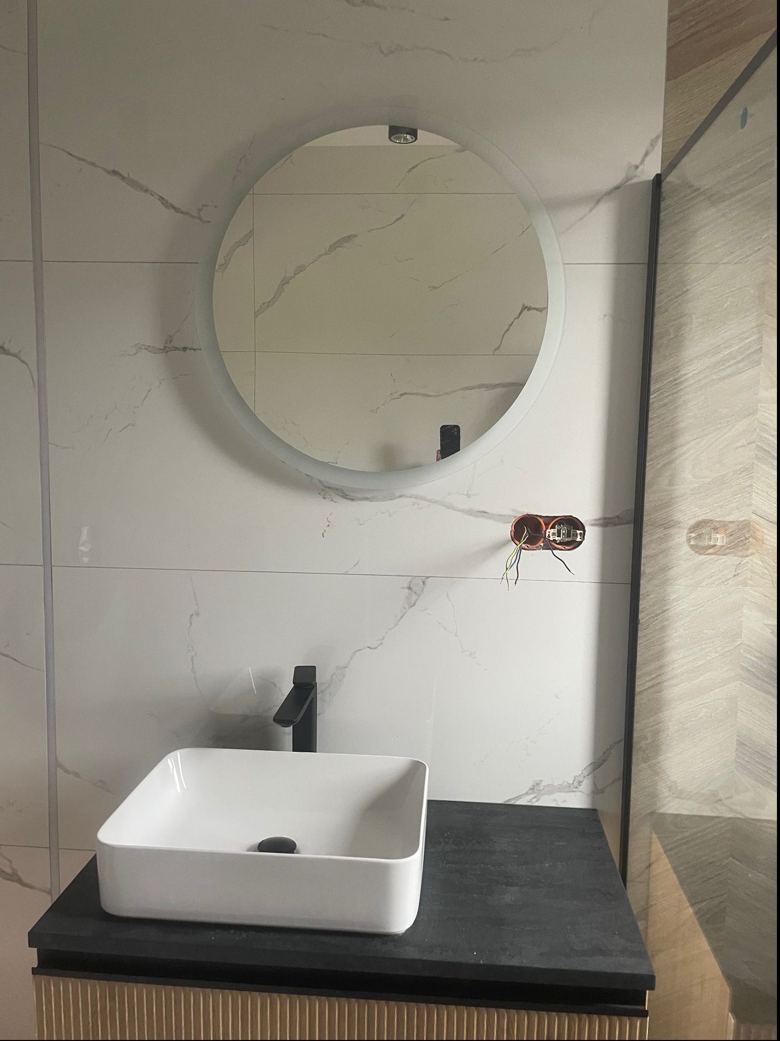 Nowoczesna łazienka w trakcie wykańczania: biała, prostokątna umywalka nablatowa z czarną armaturą, okrągłe lustro z podświetleniem na ścianie z płytek imitujących marmur, widoczne przewody...