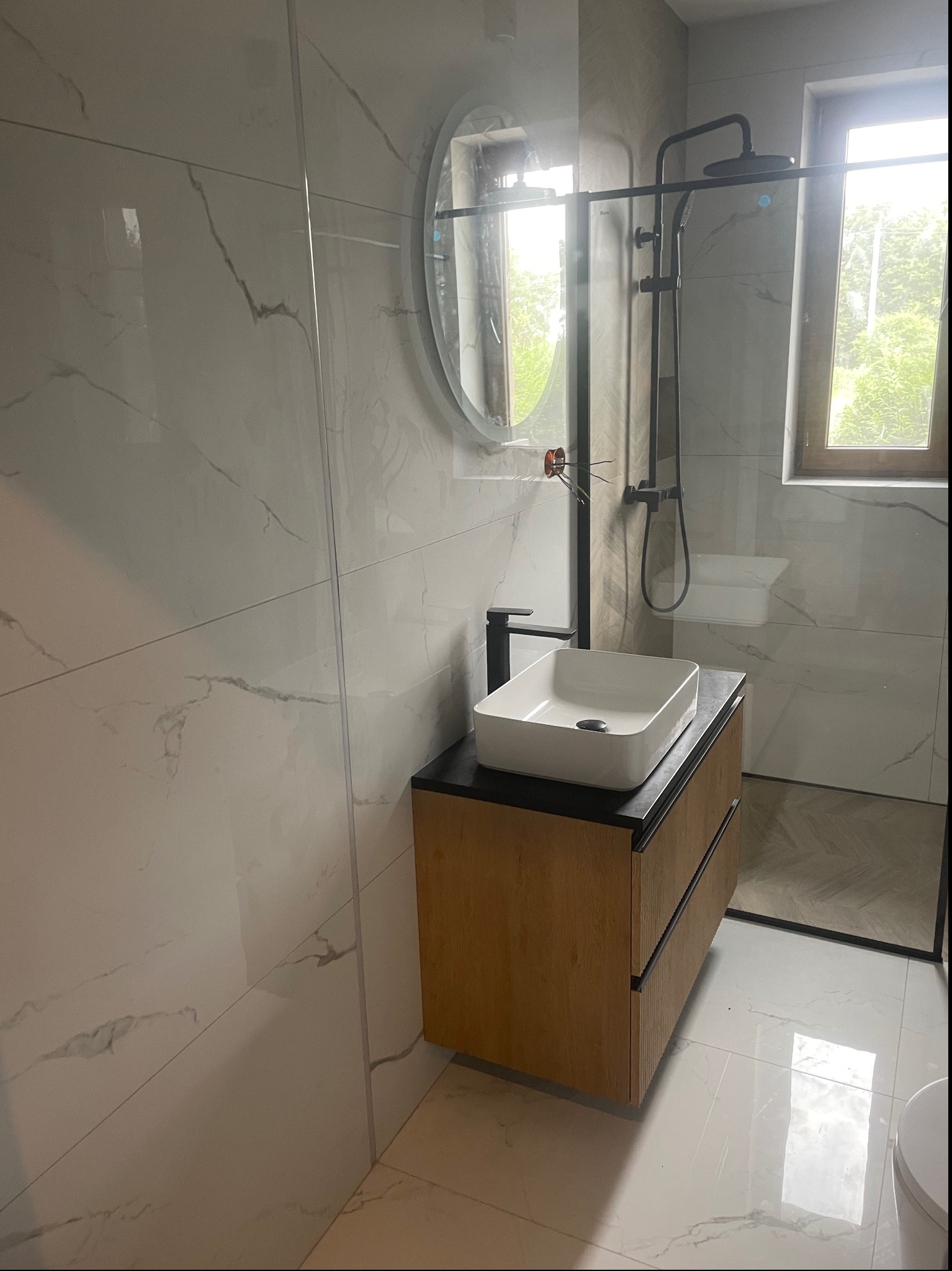 Nowoczesna łazienka z kabiną prysznicową typu walk-in, umywalką nablatową na drewnianej szafce i lustrem owalnym, wykończona białymi płytkami imitującymi marmur.