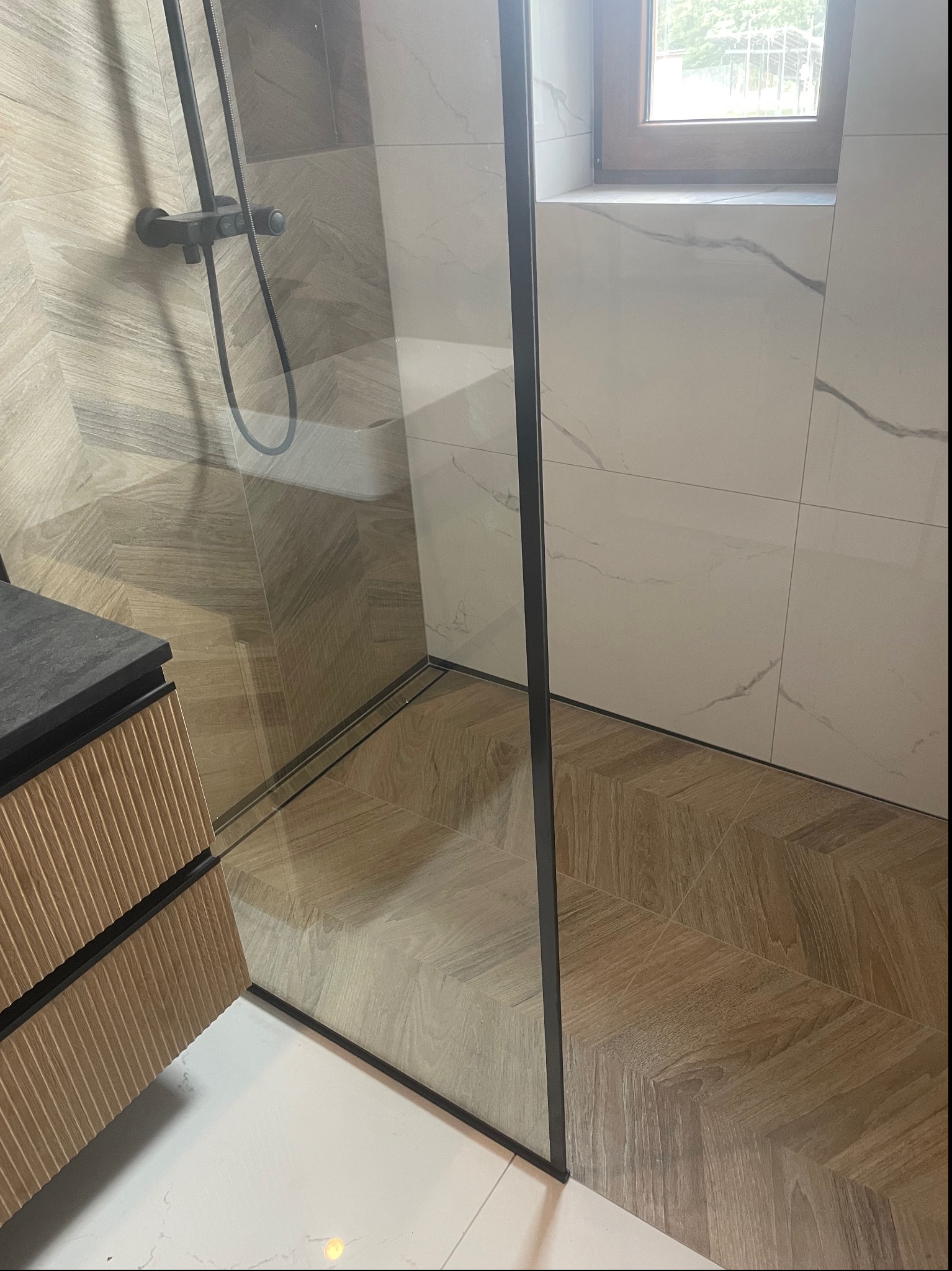 Nowoczesna kabina prysznicowa z czarną armaturą, drewnopodobną podłogą ułożoną w jodełkę i białymi płytkami imitującymi marmur na ścianach, widok z perspektywy narożnika.