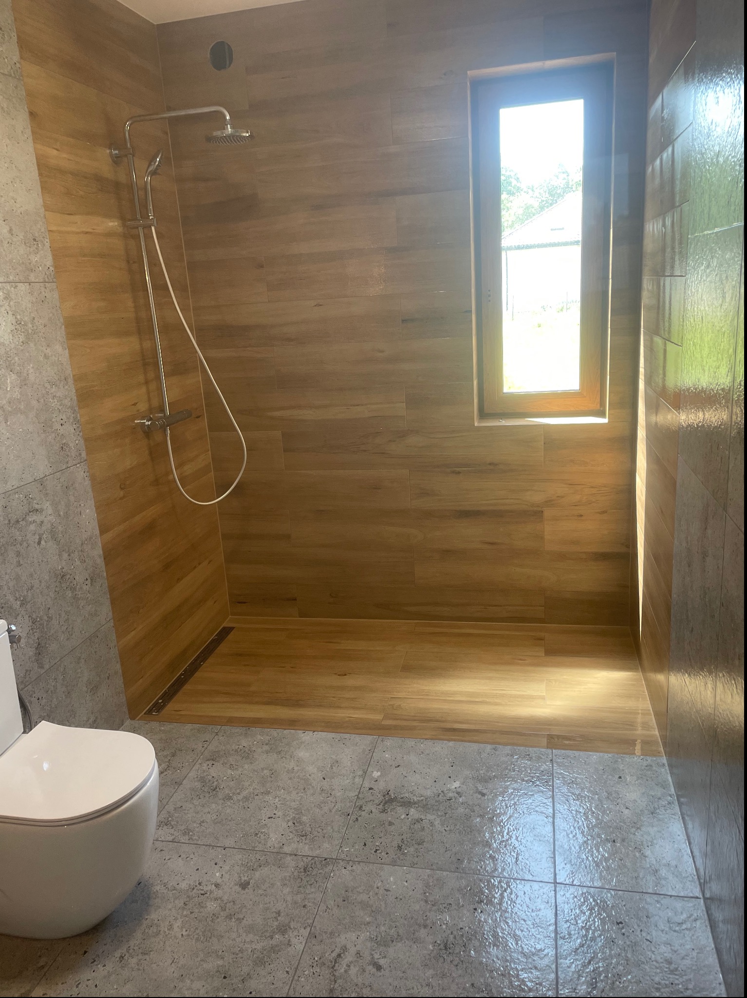 Wykończona łazienka z kabiną prysznicową typu walk-in, drewnopodobnymi płytkami ściennymi i szarymi płytkami podłogowymi, widoczne okno z naturalnym światłem.