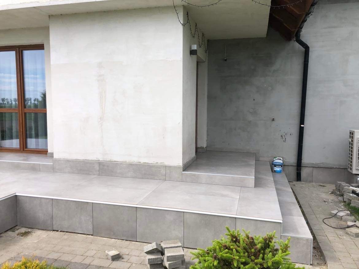 Wykończony taras z szarych płytek ceramicznych przed domem, widoczne detale wykończeniowe, fragment kostki brukowej i niewielki krzew na pierwszym planie.