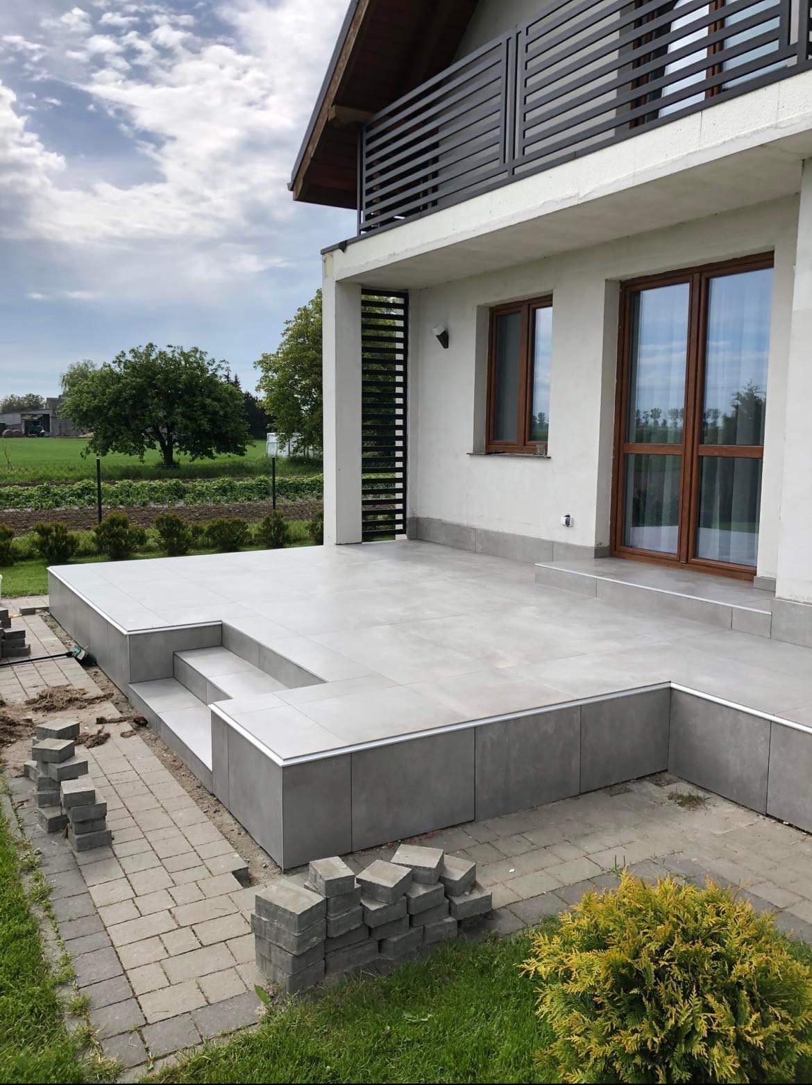 Wykończony taras z szarych płytek z niewielkimi schodami, obok sterta kostki brukowej i trawnik