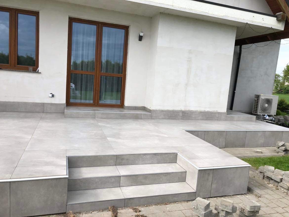 Wykończony taras z szarych płytek ceramicznych, schody z płytek, brązowe okna i drzwi tarasowe, biała elewacja budynku, klimatyzator na zewnątrz.