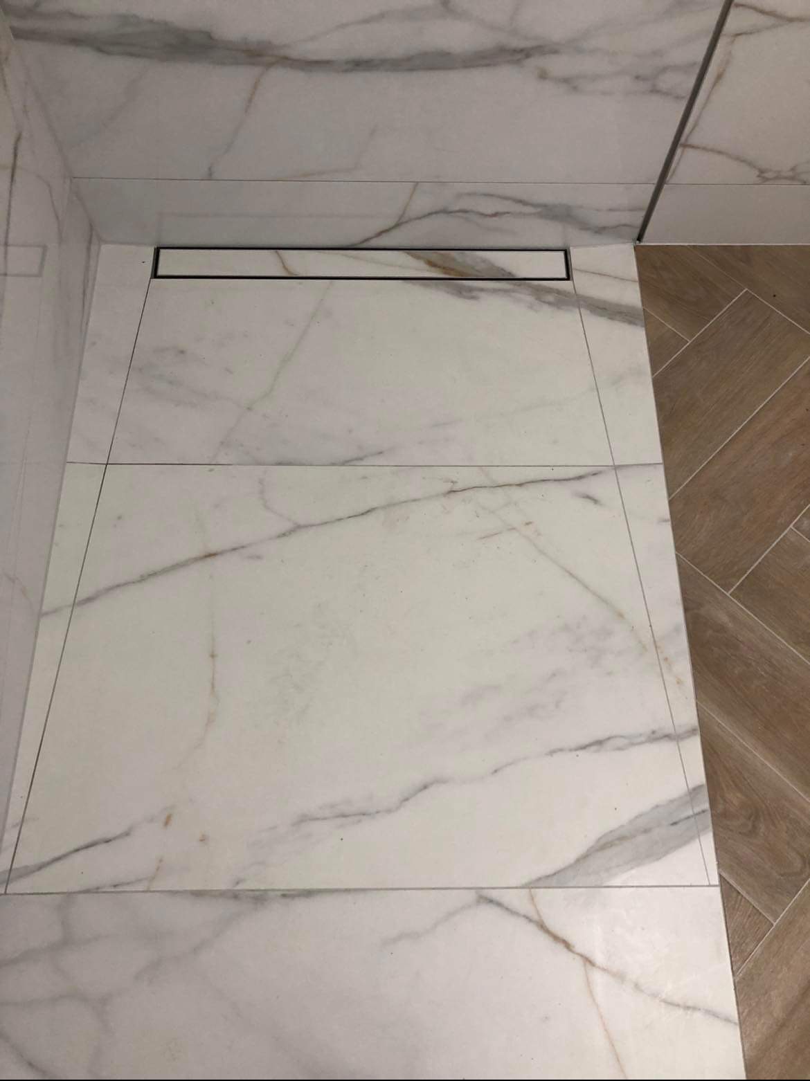 Białe, marmurowe płytki podłogowe w kabinie prysznicowej z liniowym odpływem, widok z góry, fragment drewnopodobnej podłogi w tle.