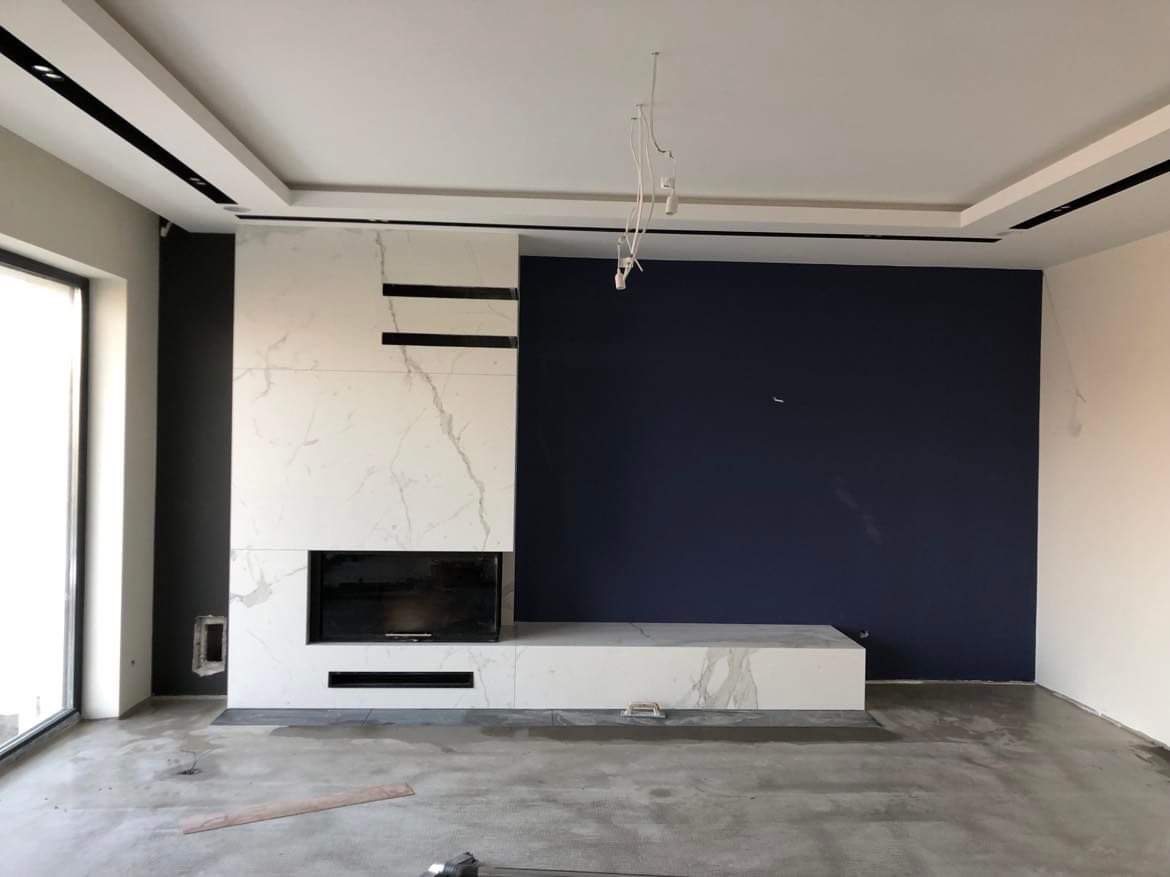 Wnętrze w trakcie prac wykończeniowych: ściana akcentowa w kolorze granatowym, zabudowa kominkowa z białego marmuru z czarnymi akcentami, surowa podłoga, widoczne niedokończone instalacje...