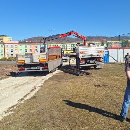 Agromania - Dwa samochody ciężarowe z dźwigiem hydraulicznym, jeden z ładunkiem kostki, drugi z prętami zbrojeniowymi, na placu budowy w słoneczny dzień.