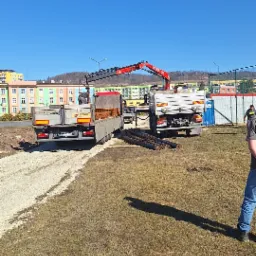 Dwa samochody ciężarowe z dźwigiem hydraulicznym, jeden z ładunkiem kostki, drugi z prętami zbrojeniowymi, na placu budowy w słoneczny dzień.