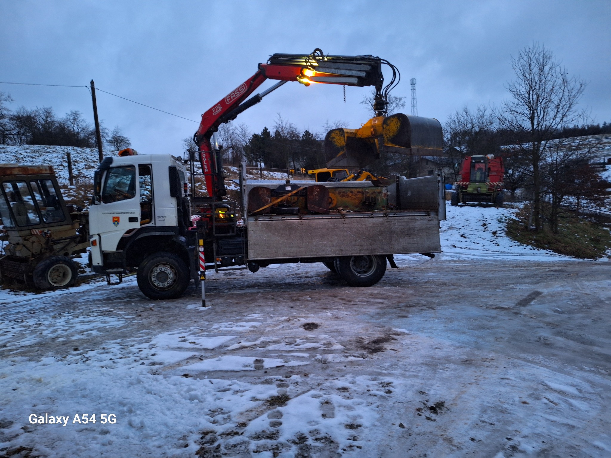 Ciężarówka z dźwigiem Fassi załadowana żółtą łyżką koparki na zasypanym śniegiem terenie budowy w pochmurny dzień.