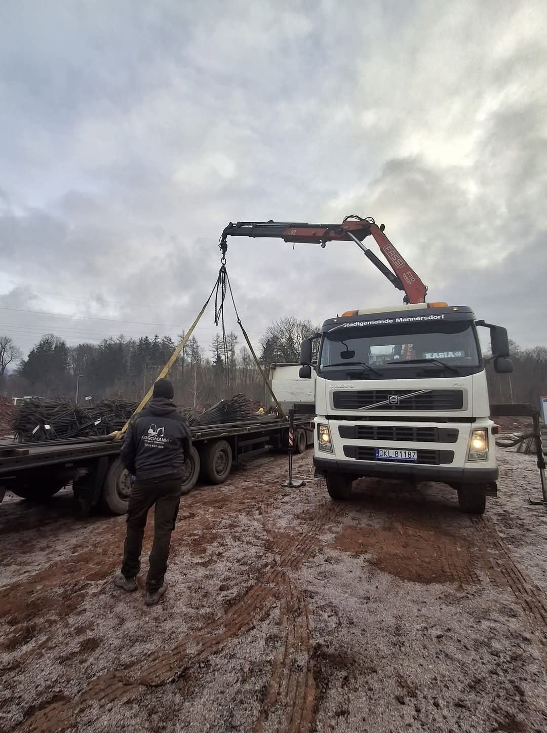 Ciężarówka z dźwigiem ładuje stalowe pręty z przyczepy na placu budowy, widoczny pracownik w kurtce z logo firmy.
