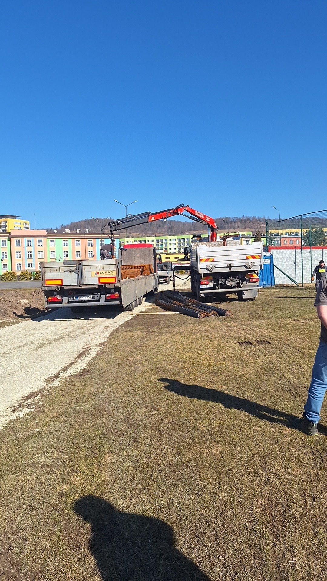 Dwa samochody ciężarowe z dźwigiem hydraulicznym, jeden z ładunkiem kostki, drugi z prętami zbrojeniowymi, na placu budowy w słoneczny dzień.