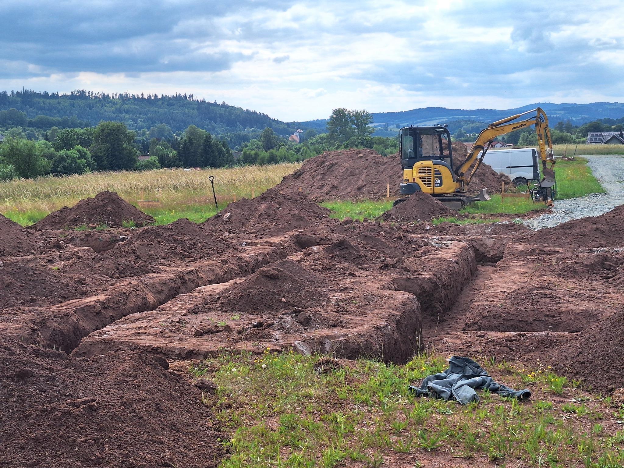 Żółta minikoparka Komatsu PC35 MR-3 przy wykopanych rowach pod fundamenty na działce z widokiem na zalesione wzgórza i zachmurzone niebo. Obok koparki stoi biały bus, a na pierwszym planie leżą...