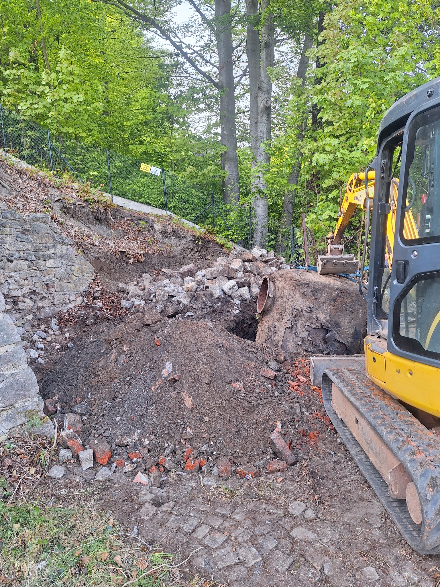Żółta mini koparka na placu budowy, wykop w ziemi z widocznym fragmentem rury, gruz i kamienie, zielone drzewa w tle.
