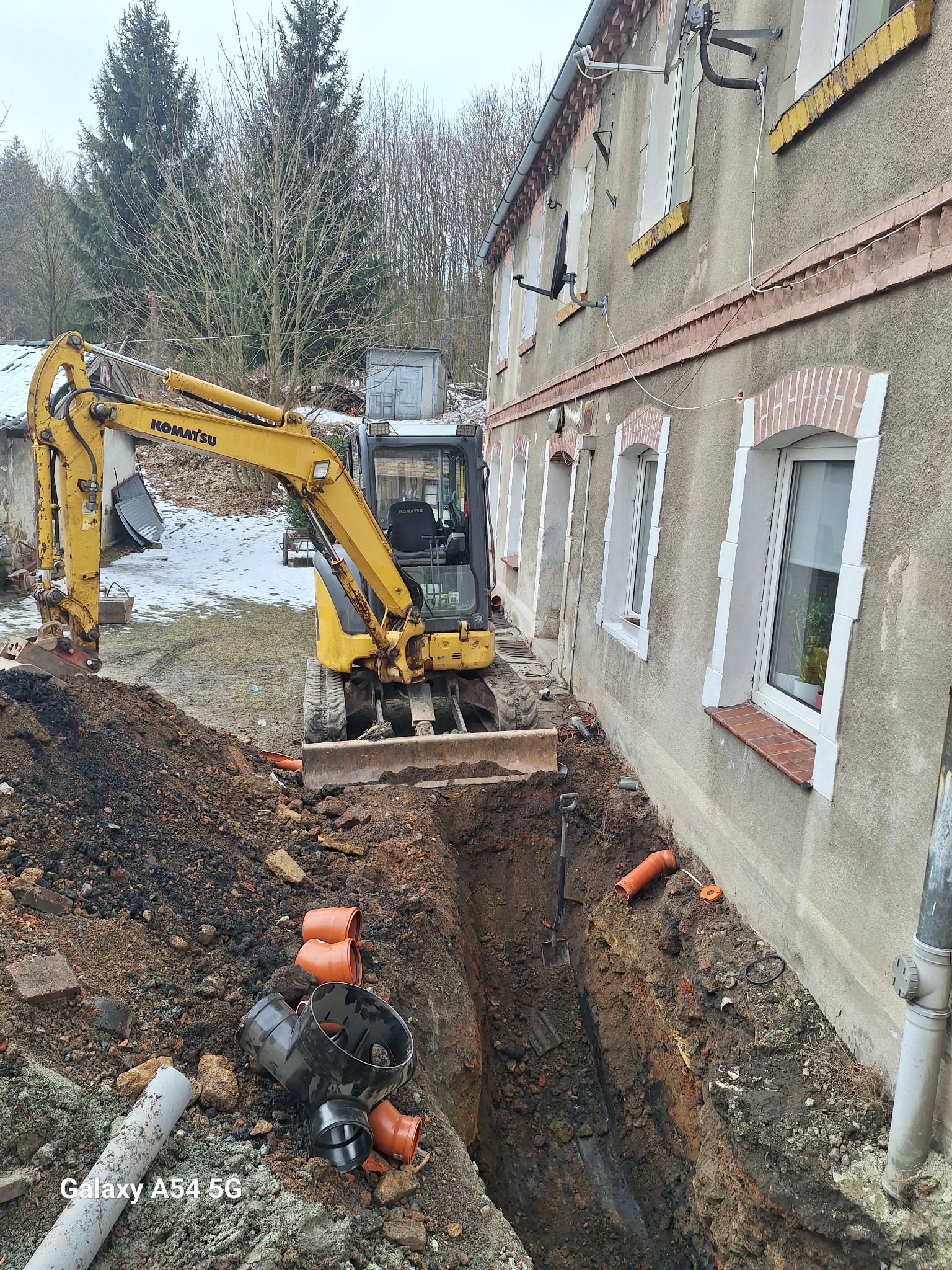 Żółta koparka Komatsu wykonuje wykop liniowy wzdłuż ściany budynku z ceglaną podmurówką, widoczne rury kanalizacyjne i osprzęt.