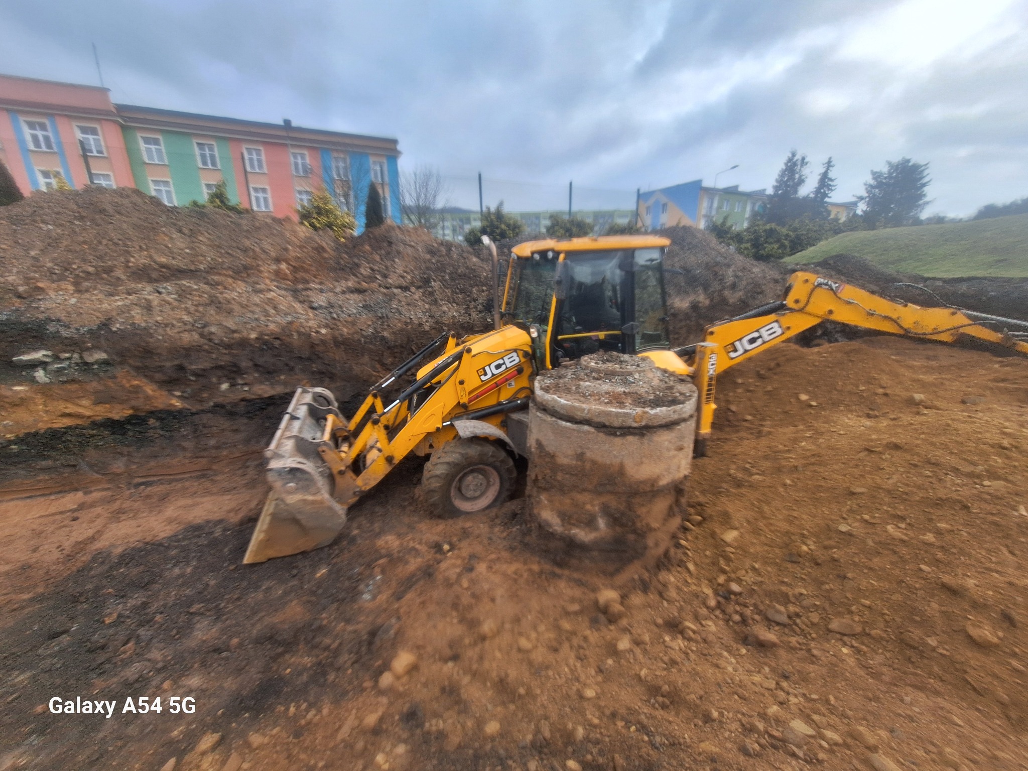 Żółta koparko-ładowarka JCB pracuje w wykopie, obok betonowy krąg studzienny, w tle kolorowy budynek i pochmurne niebo.