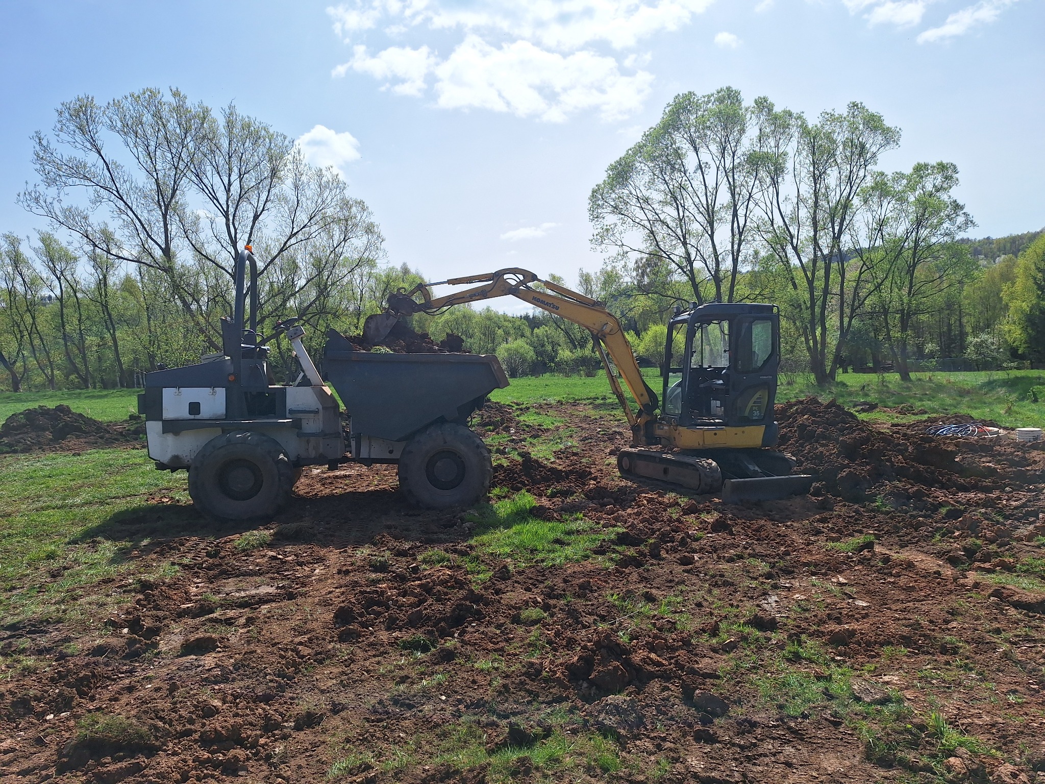 Żółta minikoparka Komatsu ładuje ziemię do wywrotki na tle zieleni, wczesna faza prac ziemnych.