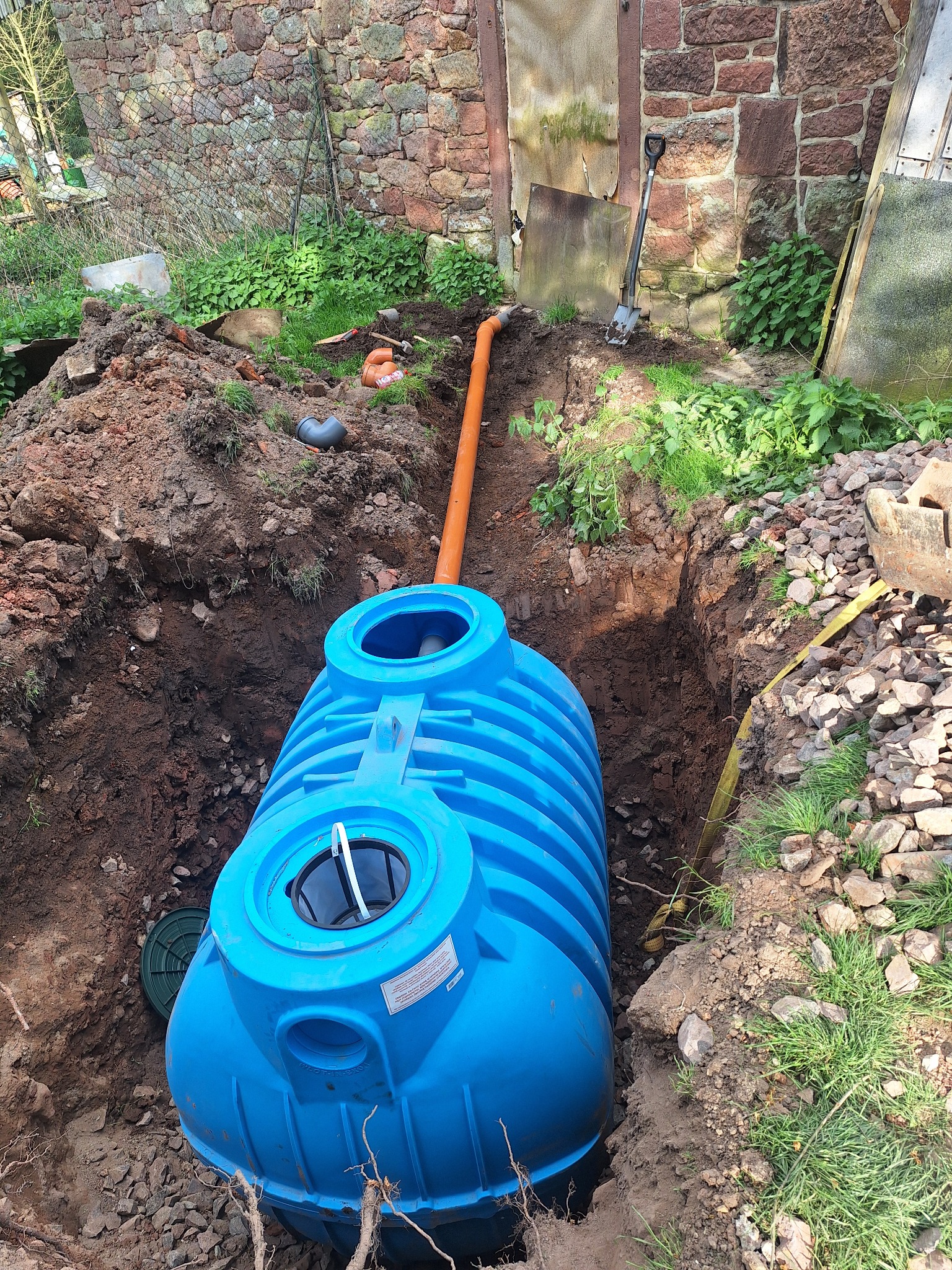 Niebieski zbiornik retencyjny częściowo zagłębiony w wykopie ziemnym, z widocznymi rurami doprowadzającymi, otoczony ziemią, kamieniami i zielenią, w tle kamienny mur i drewniane drzwi.