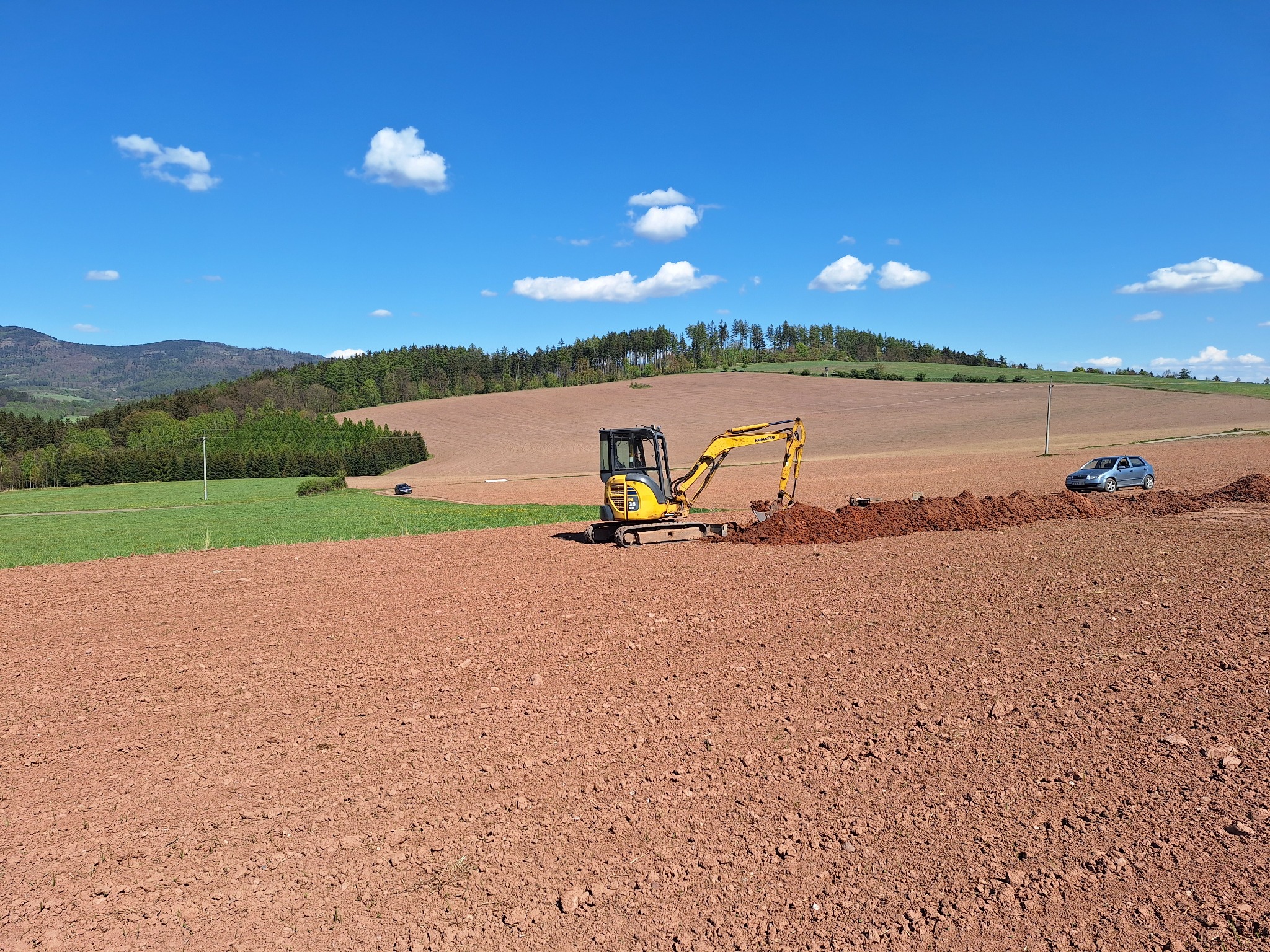 Żółta mini koparka Komatsu wykonuje wykop w polu o czerwonej glebie, na horyzoncie widoczne wzgórza i niebieskie niebo z chmurami, obok wykopu zaparkowany srebrny samochód.