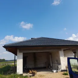 Dom w trakcie budowy z nowym, czarnym dachem z dachówki ceramicznej, widoczne białe bloczki konstrukcyjne i niebieski samochód dostawczy w tle.