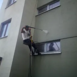 Pracownik na linach myje elewację budynku za pomocą myjki ciśnieniowej, w oknie odbija się sylwetka osoby obserwującej pracę.