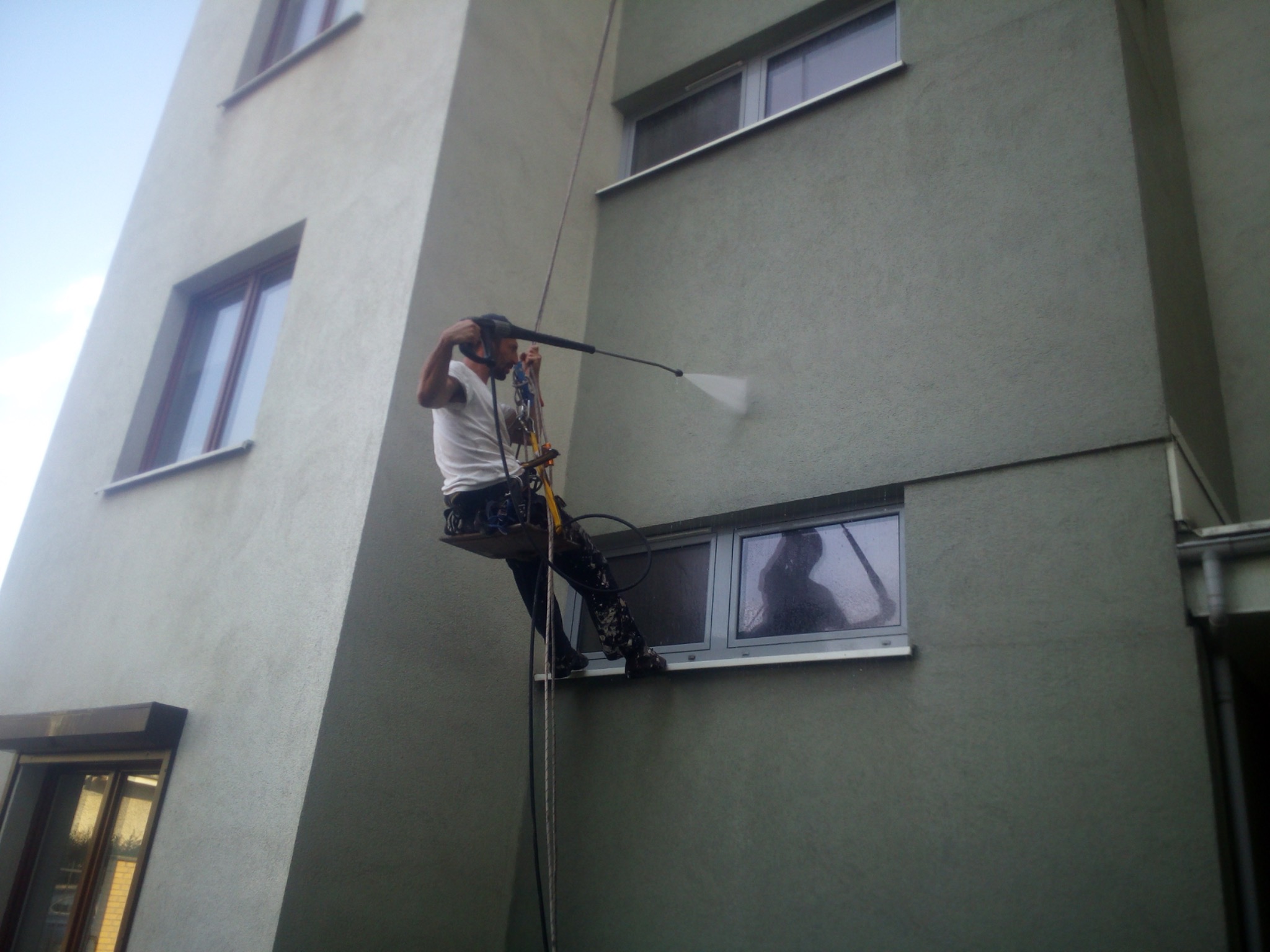 Pracownik na linach myje elewację budynku za pomocą myjki ciśnieniowej, w oknie odbija się sylwetka osoby obserwującej pracę.