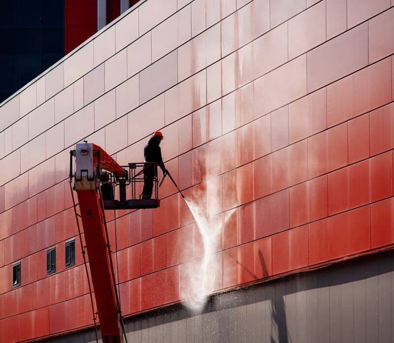 Pracownik w pomarańczowym kasku na podnośniku koszowym czyści czerwoną elewację budynku za pomocą myjki ciśnieniowej, strumień wody spływa po panelach.