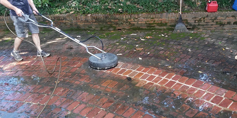 Czyszczenie czerwonej kostki brukowej za pomocą myjki ciśnieniowej z okrągłą dyszą. Widoczna różnica między czystą a brudną powierzchnią, mokre liście i resztki na kostce.