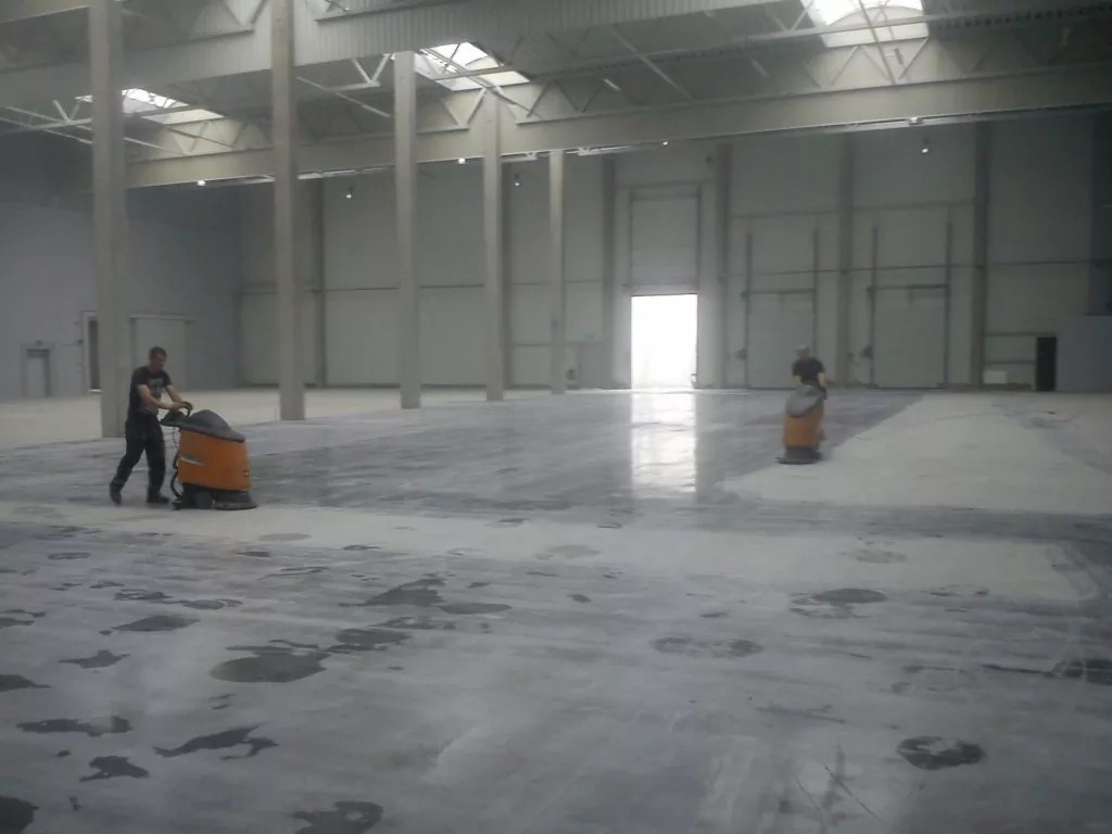 Pracownik obsługuje pomarańczową maszynę do czyszczenia posadzek w przestronnej hali magazynowej z betonową podłogą, widoczne jasne plamy po myciu i wysokie słupy konstrukcyjne.