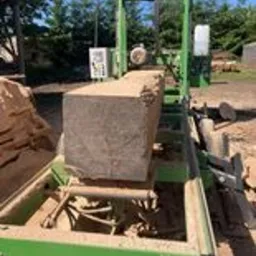 Blok drewna umieszczony na zielonej poziomej pile taśmowej, przygotowany do cięcia, z widocznymi trocinami i elementami sterującymi maszyną w tle.