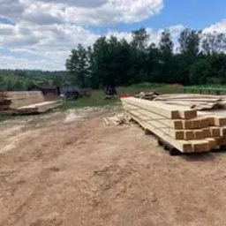 Widok placu składowego w Strzegomiu z ułożonymi na paletach kantówkami oraz stosem desek w tle, z zielenią drzew i błękitnym niebem z chmurami.