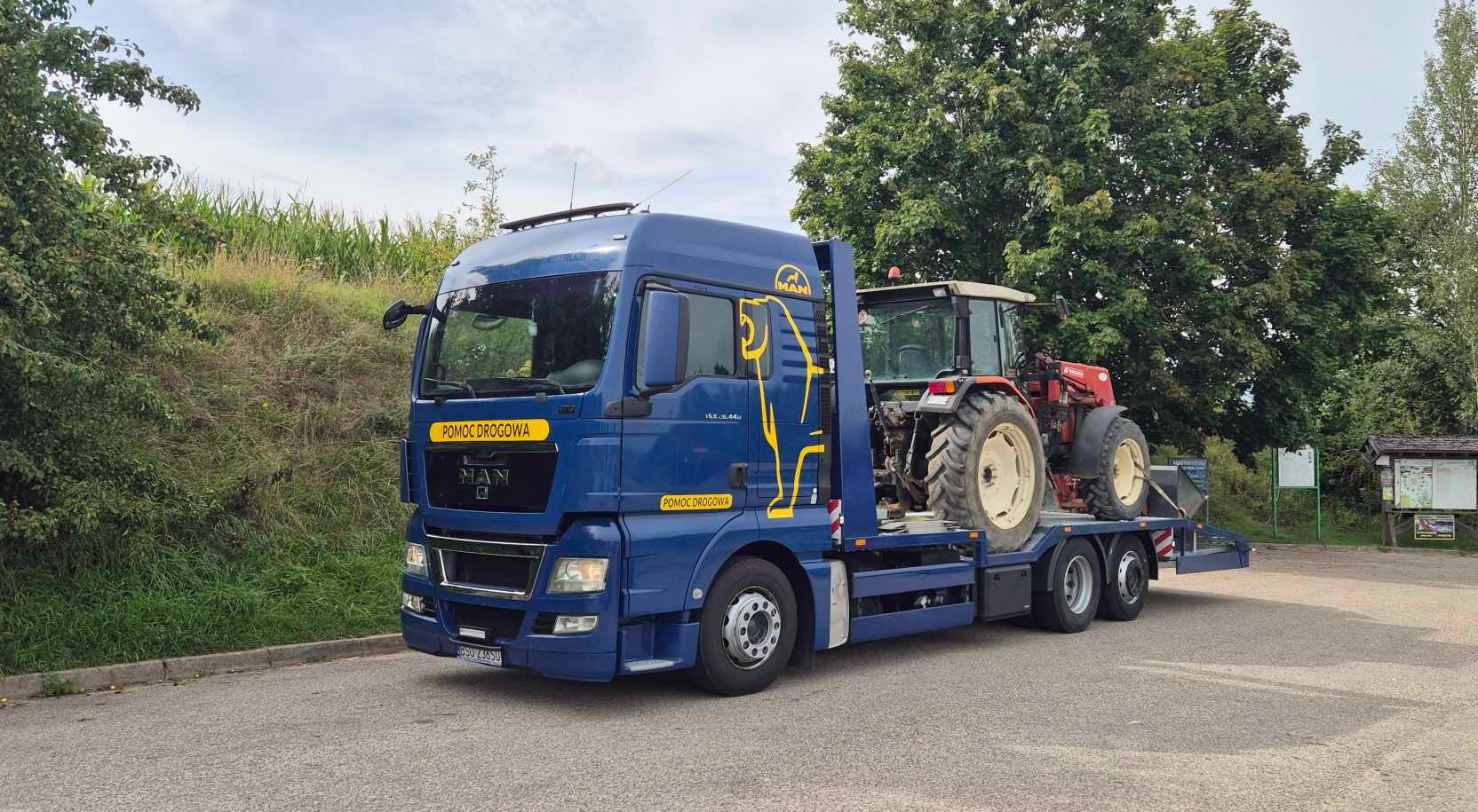 Niebieska ciężarówka pomocy drogowej MAN z żółtym logo dźwigu przewozi czerwony traktor na platformie, widok z boku na tle zieleni.